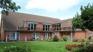 Une maison à deux étages, construite en briques, dotée d’un patio couvert, d’un petit jardin avec diverses plantes et fleurs, et surplombant un ciel dégagé.