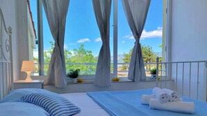 A bedroom with a bed, a window with curtains, a view of greenery, and a balcony with a railing.