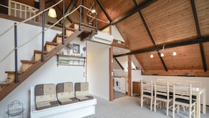 Un loft amplio con techo alto, vigas de madera a la vista, una escalera, una zona de comedor con sillas blancas y un área de lavandería.