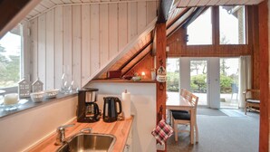 Una cocina con encimera de madera, un lavamanos y una cafetera. Hay una zona de comedor con mesa y sillas, y un ventanal con vistas al exterior.