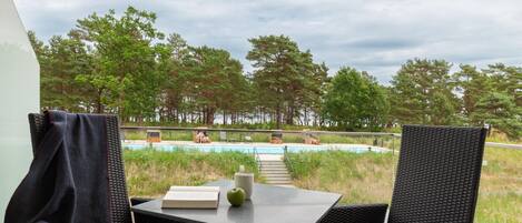 A balcony with wicker chairs and a table, overlooking a swimming pool and a forested area.