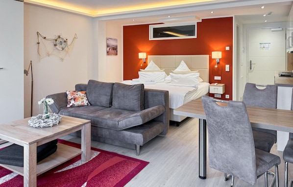 A modern hotel room with a grey sofa, a wooden coffee table, a red and white rug, a bed with white linens, and a small desk with a chair. - Cozy apartment in Binz with house sea view (Prora/Rügen)