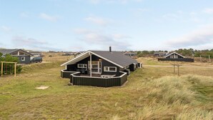 A modern house with a large deck, surrounded by a grassy field and other houses in the background.