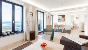 Una sala de estar moderna con comedor, ventanas amplias con cortinas y vista al océano, junto con un rincón de descanso acogedor.