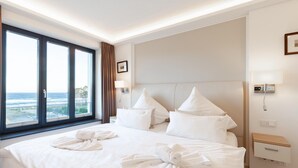 Quarto de hotel com uma cama grande, duas almofadas, uma mesa de cabeceira, um abajur e vista para o oceano através de uma janela ampla.
