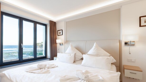 Habitación de hotel con una cama grande, dos almohadas, una mesita de noche, una lámpara y vistas al océano a través de un ventanal amplio.