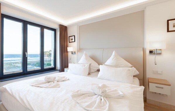 A hotel room with a large bed, two pillows, a nightstand, a lamp, and a view of the ocean through a large window.