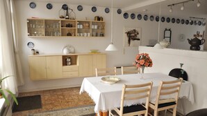 Uma área de jantar com uma mesa posta para dois, cadeiras de madeira, um vaso de flores e um armário com copos.
