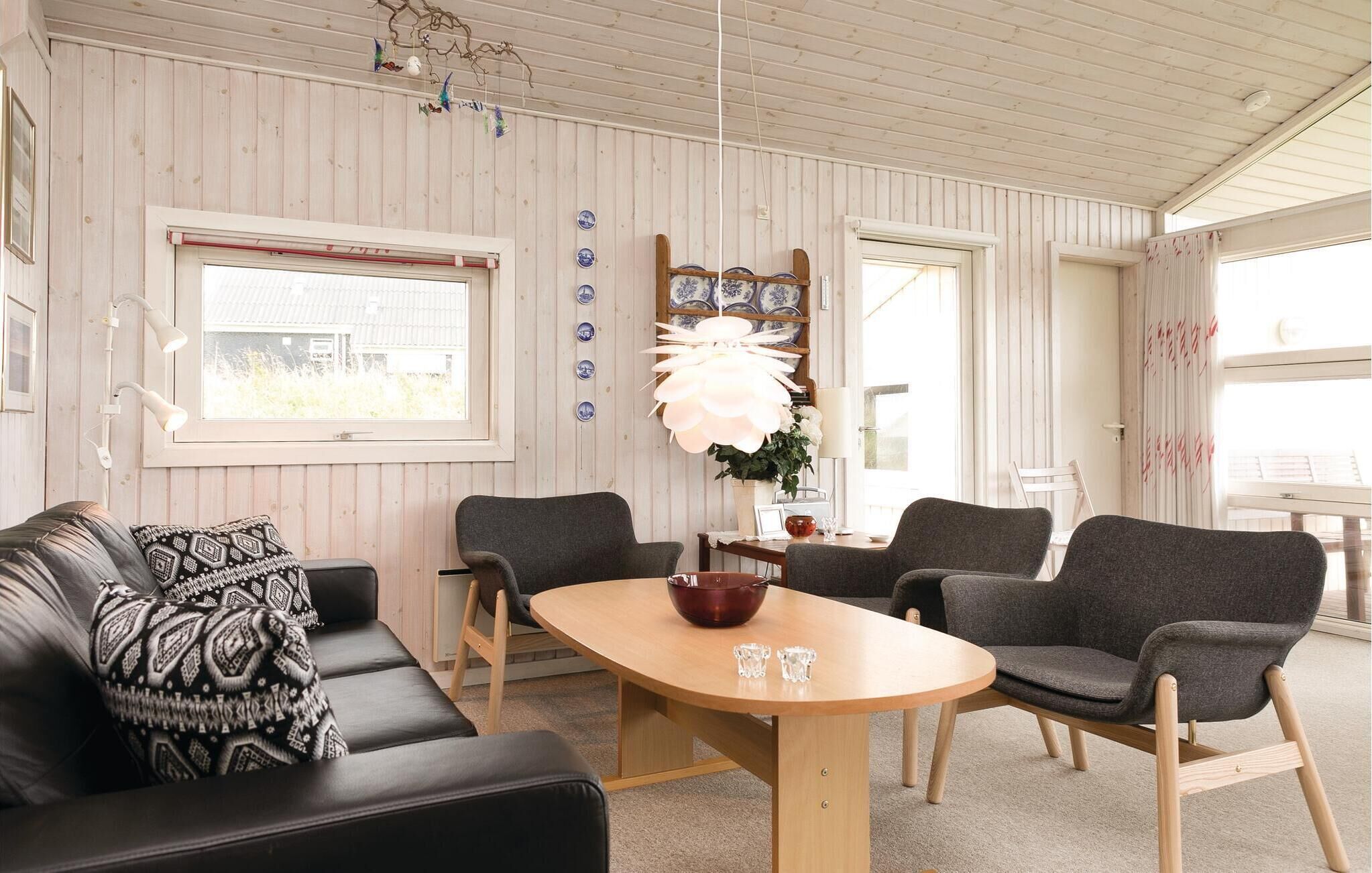 A cozy living room with a wooden ceiling, a black leather sofa with patterned cushions, a wooden coffee table, and grey armchairs.