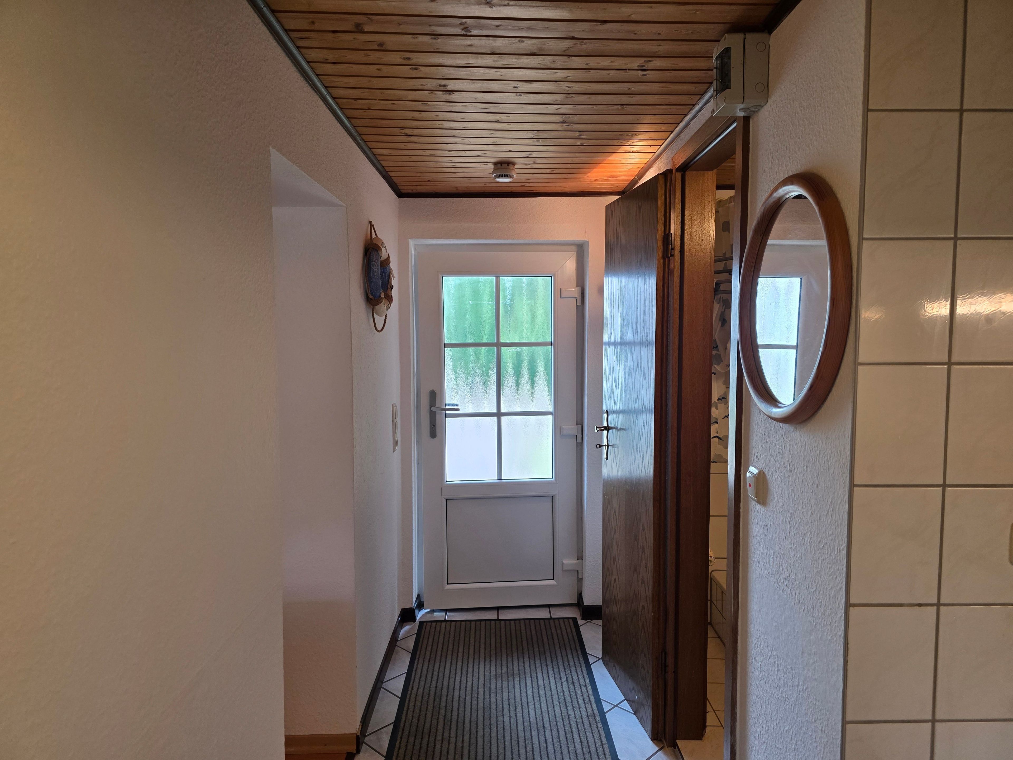 A hallway with a wooden ceiling, a door with a frosted glass window, a mat at the entrance, and a wall-mounted mirror.