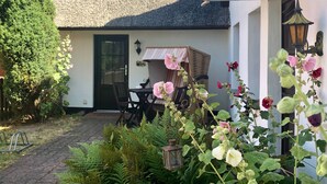 Une maison avec un toit de chaume, une porte, une table avec des chaises et un jardin agrémenté de fougères et de fleurs.