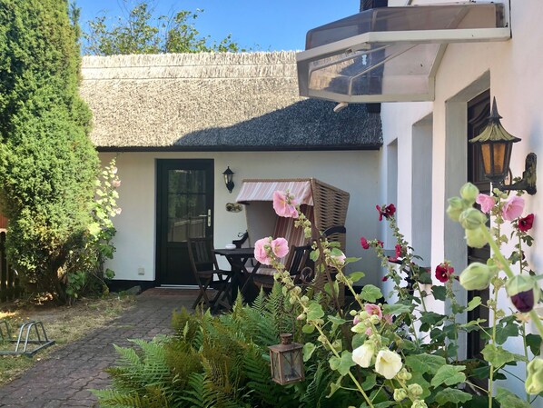 Ein Haus mit einem Strohdach, einer Tür, einem Tisch mit Stühlen und einem Garten mit Farne und Blumen.