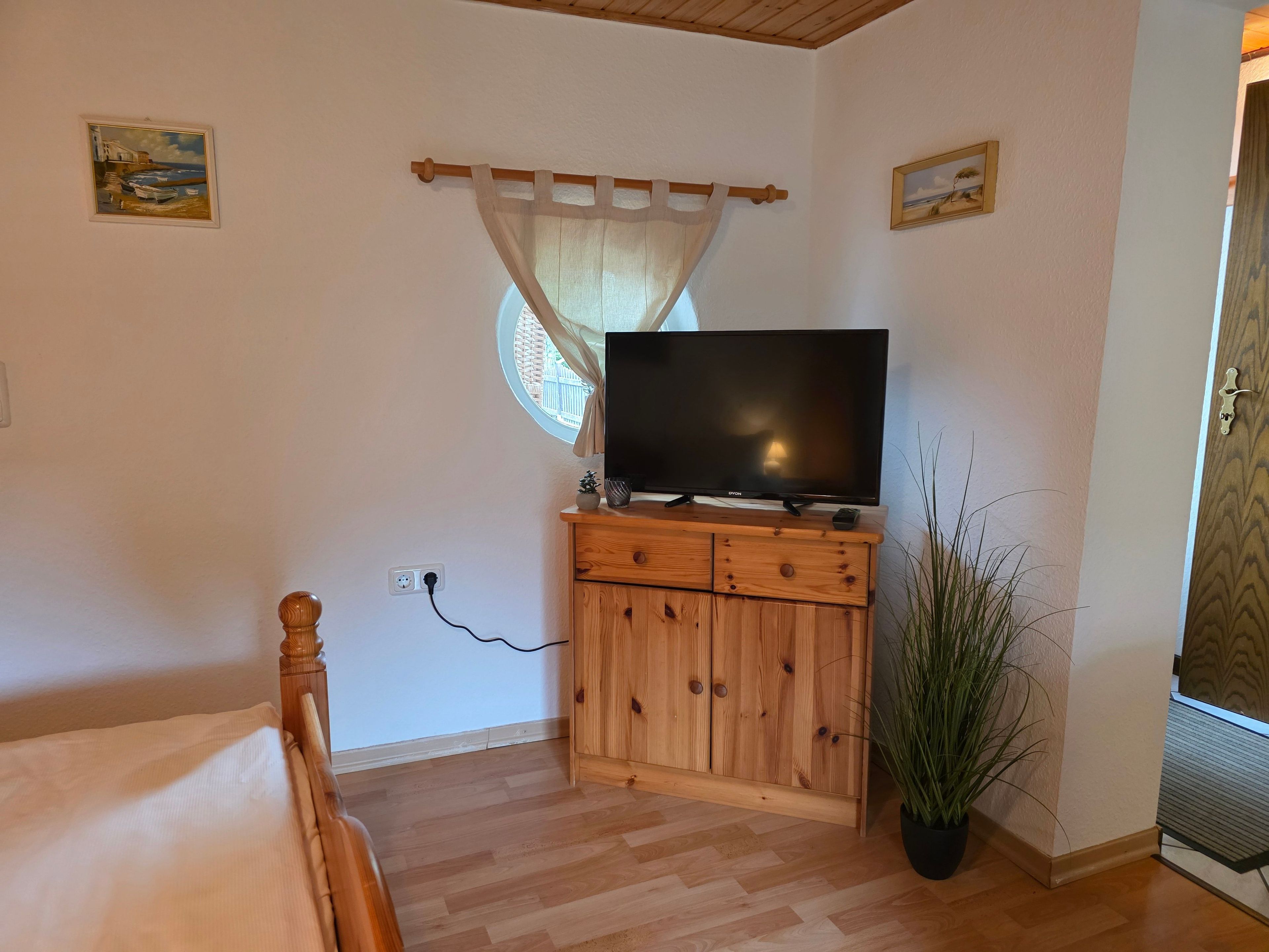A bedroom with a bed, a wooden cabinet with a flat-screen TV, a window with a curtain, and a potted plant.