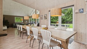 Un comedor amplio con una larga mesa de madera y sillas blancas, ventanas grandes que ofrecen vistas a zonas verdes, y una cocina moderna al fondo.