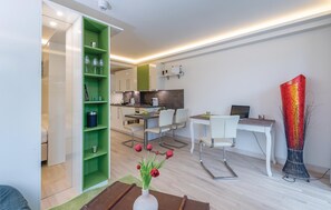 A modern living room with a green bookshelf, a dining area with white chairs, a kitchen in the background, and a red vase on a table.