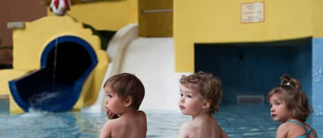 Niños en una piscina con un tobogán acuático y un edificio con ventanas arqueadas al fondo.