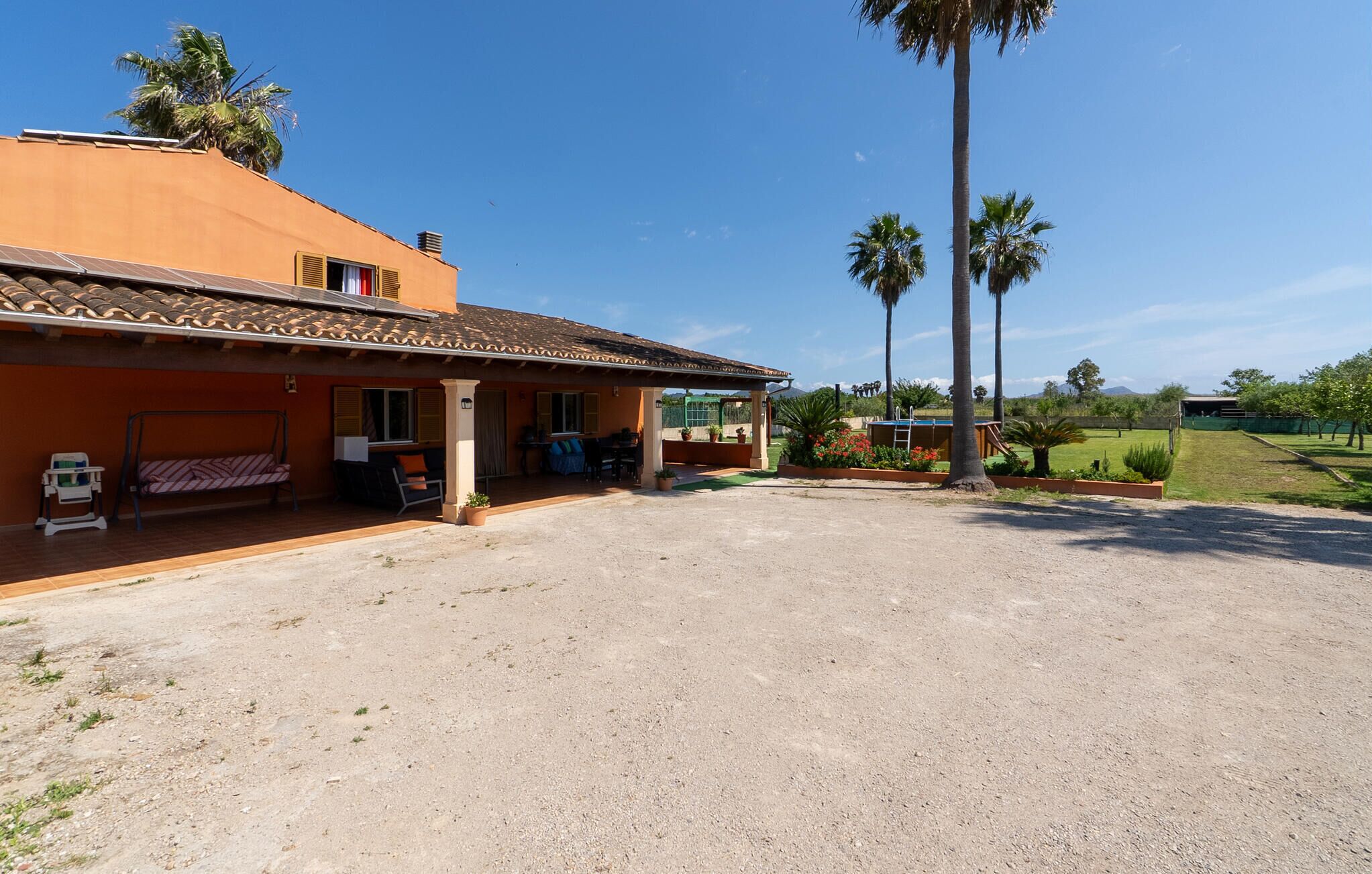 Un bâtiment à deux étages doté d’un patio, d’un espace extérieur couvert avec des sièges, et offrant une vue sur un champ verdoyant et des palmiers.