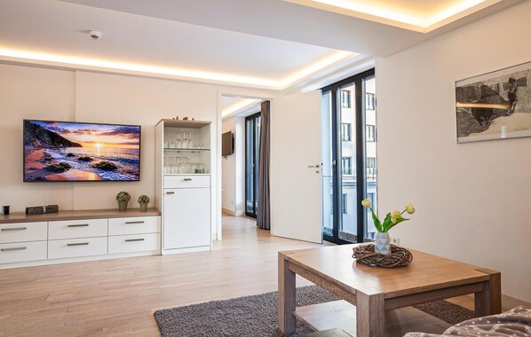 A modern living room with a flat-screen TV, a wooden coffee table, and a view of the city through large windows.