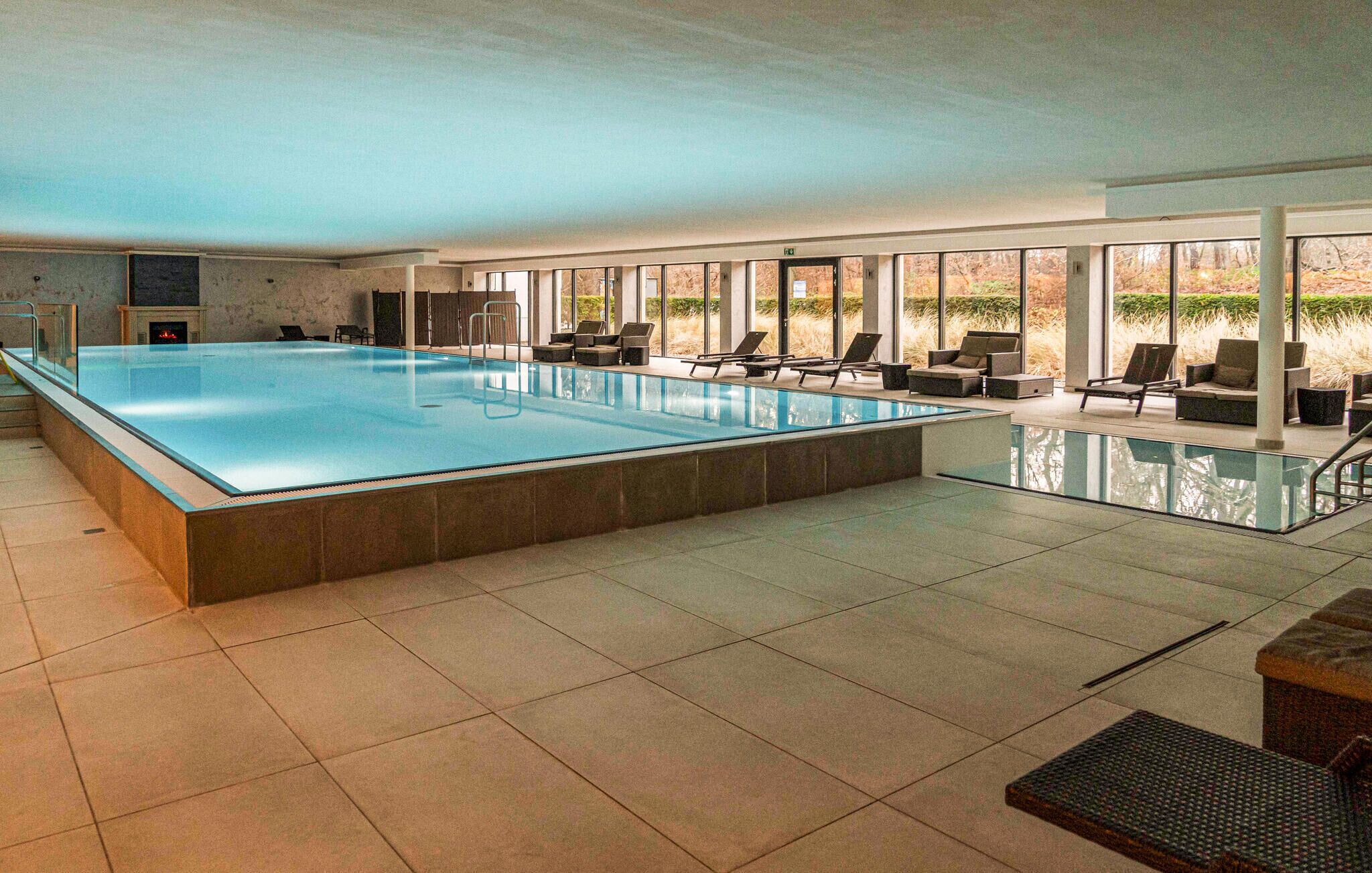 A large indoor swimming pool with a tiled floor, surrounded by lounge chairs and a glass railing.
