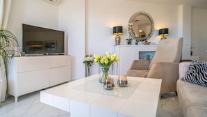 A modern living room with a flat-screen TV, a glass coffee table, a beige sofa, and a large mirror.