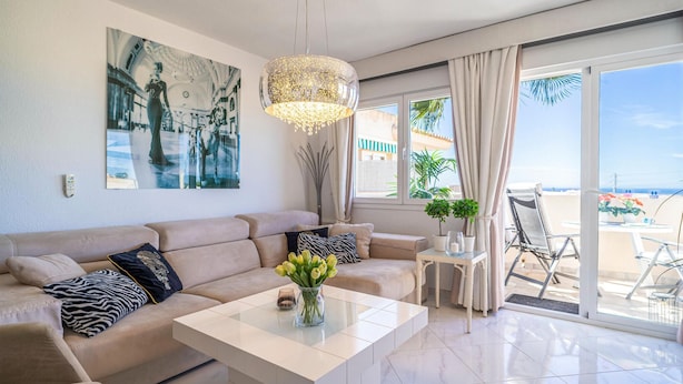 A modern living room with a beige sofa, a glass coffee table, and a chandelier. There is a large window with curtains, a balcony with a chair, and a painting on the wall.