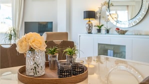 A dining area with a table set for two, featuring a vase with flowers, potted plants, and candle holders.