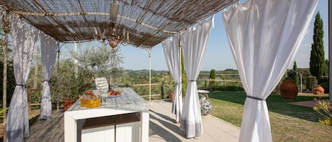 Une terrasse extérieure avec une table dressée pour le repas, entourée de rideaux blancs et surmontée d’un auvent en bambou.