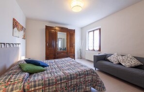 Ein Schlafzimmer mit einem Bett, einem grauen Sofa, einem Holzschrank, einem Fenster mit Vorhängen und einer Deckenleuchte.