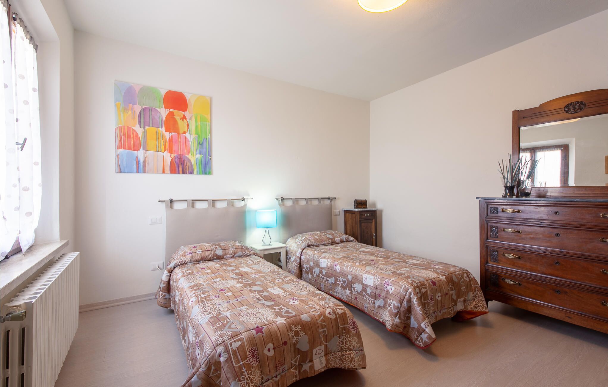 A bedroom with two single beds, a dresser, a radiator, a wall-mounted lamp, and a colorful abstract painting.