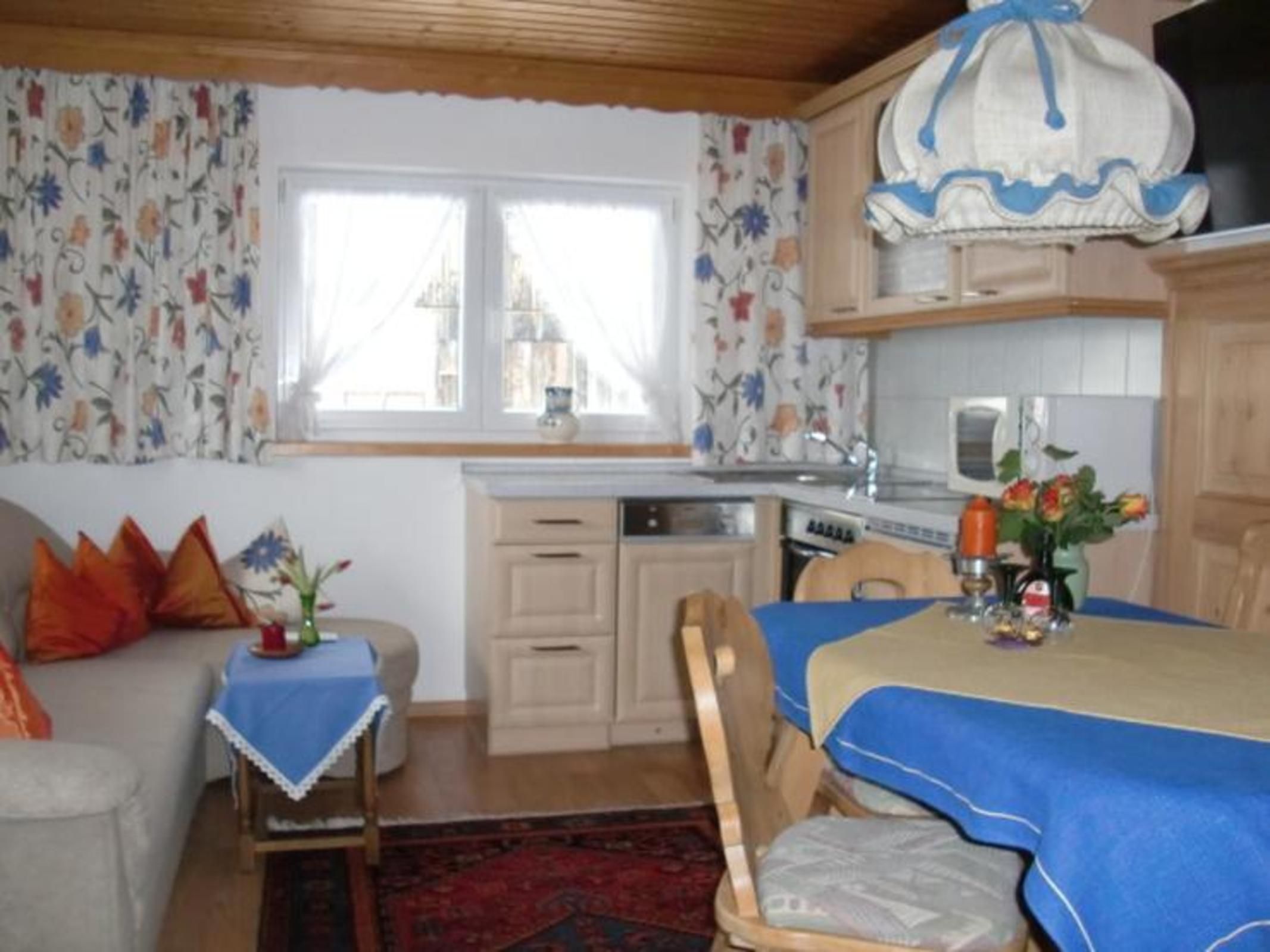 Ein gemütliches Zimmer mit Esstisch, Sofa, einem Fenster mit Vorhängen sowie einer Küchenzeile mit Schränken und einer Mikrowelle.