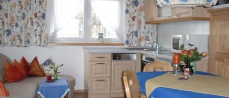 Ein gemütliches Zimmer mit Esstisch, Sofa, einem Fenster mit Vorhängen sowie einer Küchenzeile mit Schränken und einer Mikrowelle.