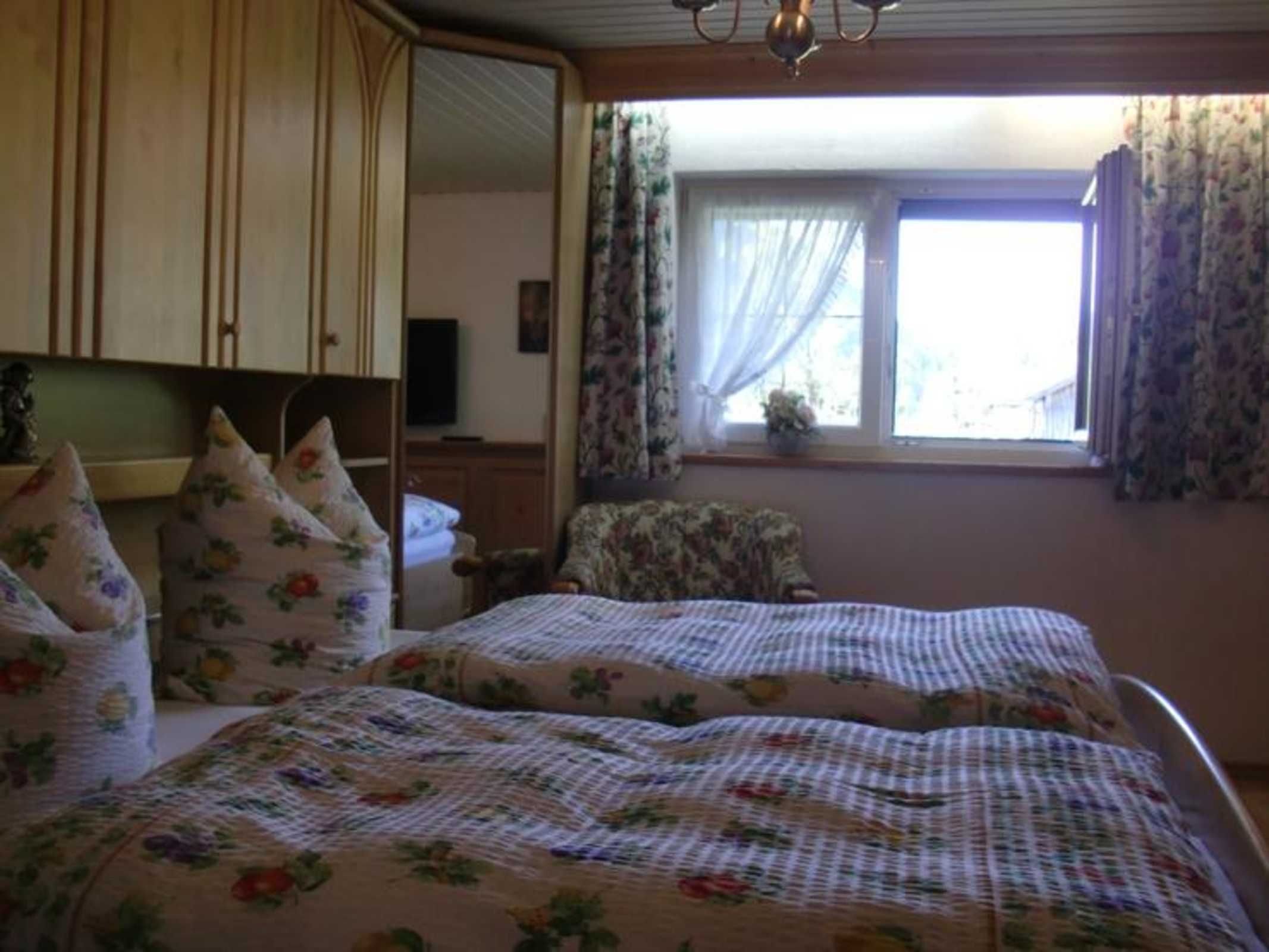 Un dormitorio con dos camas, ropa de cama estampada con motivos florales, una silla, un televisor y un ventanal con cortinas.