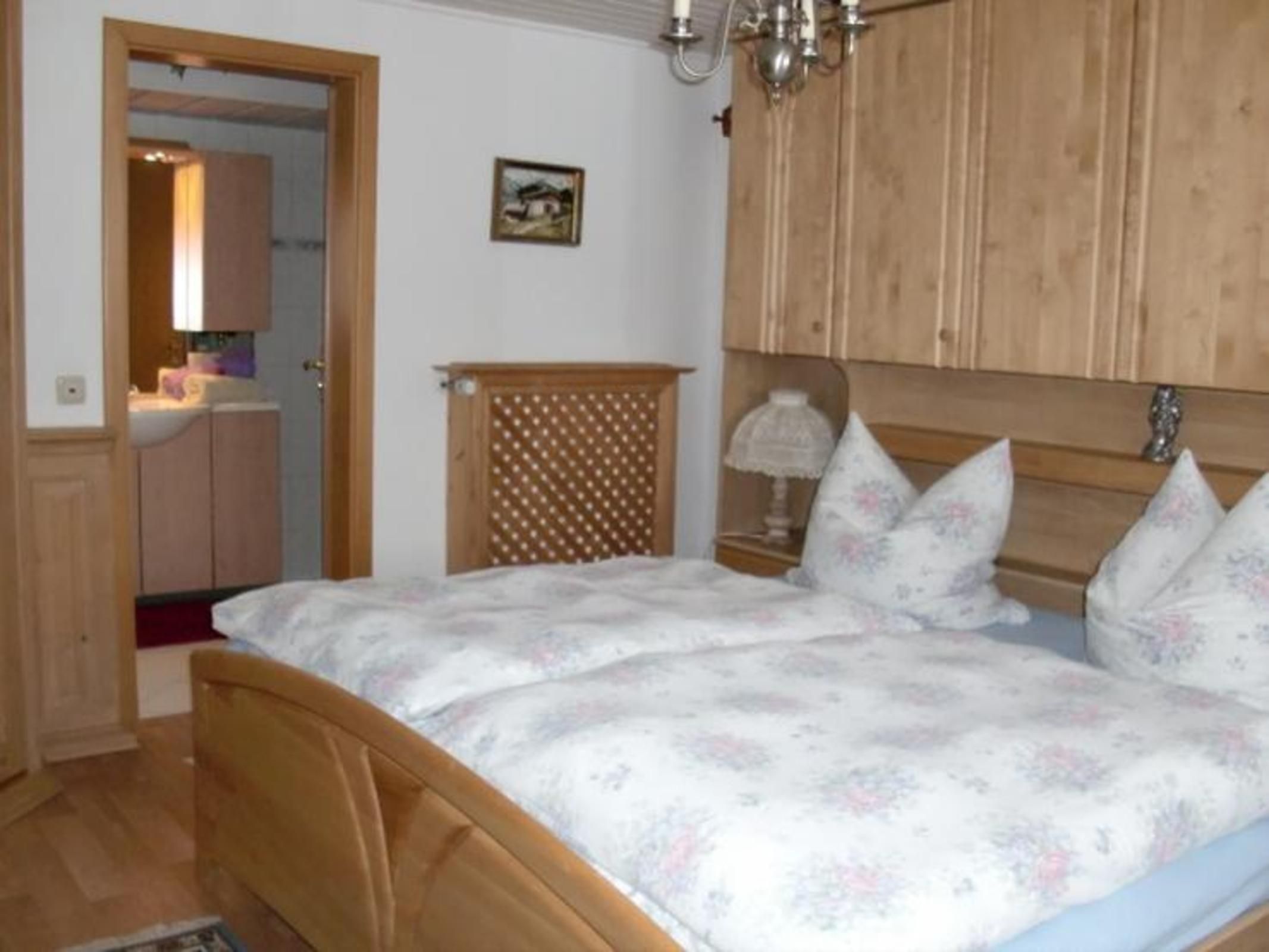 A bedroom with a wooden bed, a nightstand with a lamp, a wooden wardrobe, and a small painting on the wall.