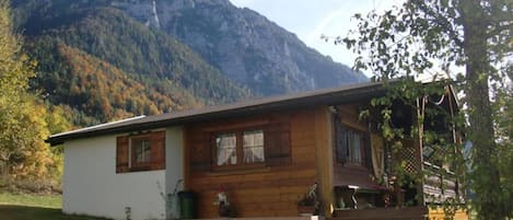 A wooden cabin with a porch, a small garden, and a mountain backdrop.