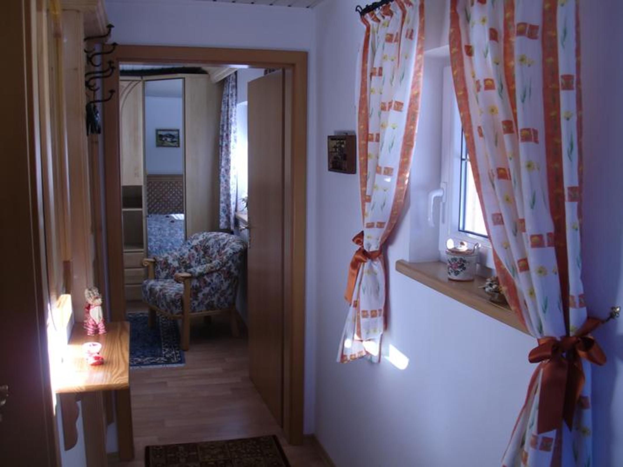 Un pasillo con una puerta de madera, una silla, una mesita con una lámpara y una ventana con cortinas.