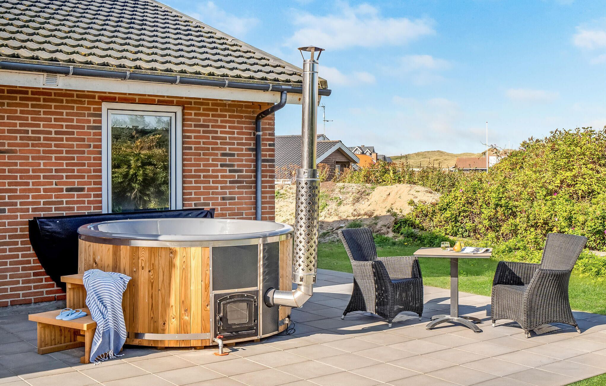 A hot tub with a wooden exterior, a brick house with a tiled roof, and outdoor wicker chairs.