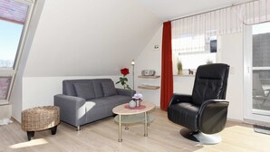 Ein modernes Wohnzimmer mit einem grauen Sofa, einem schwarzen Ledersessel, einem Korbstuhl und einem kleinen runden Tisch mit einer Vase und einer Lampe.