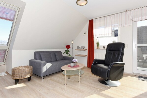 A modern living room with a grey sofa, a black leather chair, a wicker stool, and a small round table with a vase and a lamp.