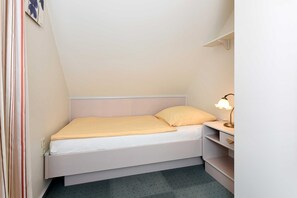 A small, single-bed room with a white headboard, a bedside table with a lamp, and a shelf unit.