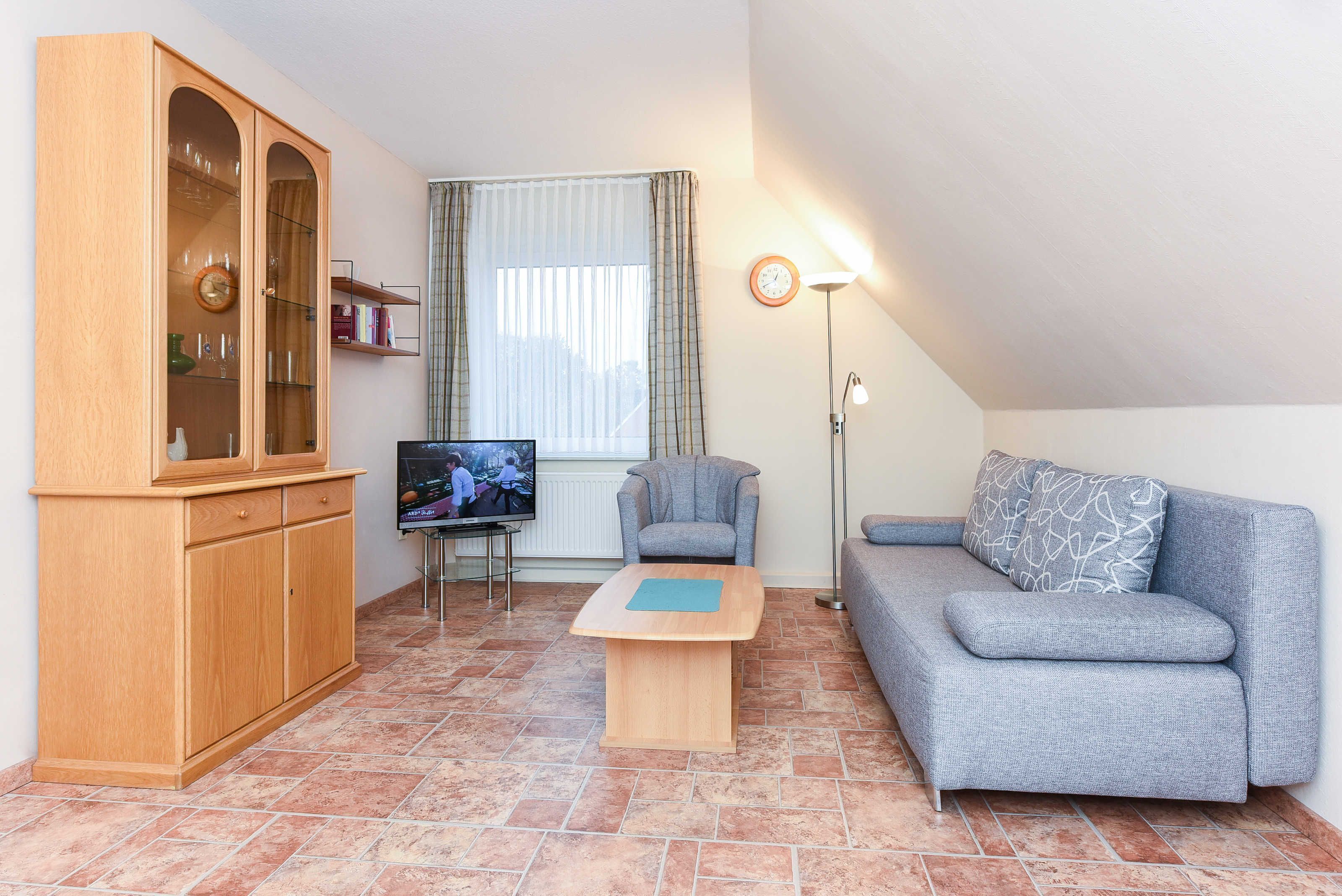 Een woonkamer met een houten kast, een grijze bank, een kleine tafel, een televisie en een raam met gordijnen.