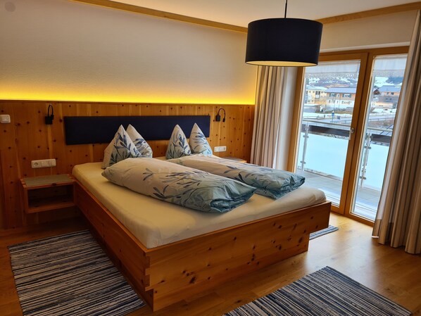 A wooden bed with leaf-patterned pillows, a bedside table, a nightstand, a black hanging lamp, and a view of a snowy landscape through a glass door.