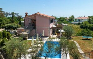 A two-story house with a red-tiled roof, a swimming pool, and a garden with various plants and a pergola.