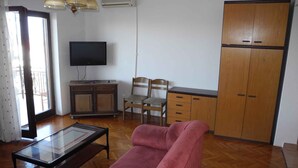 Ein Wohnzimmer mit einer Couch, einem Glastisch, einem Fernseher, einer Holzkassette und einem Sessel.