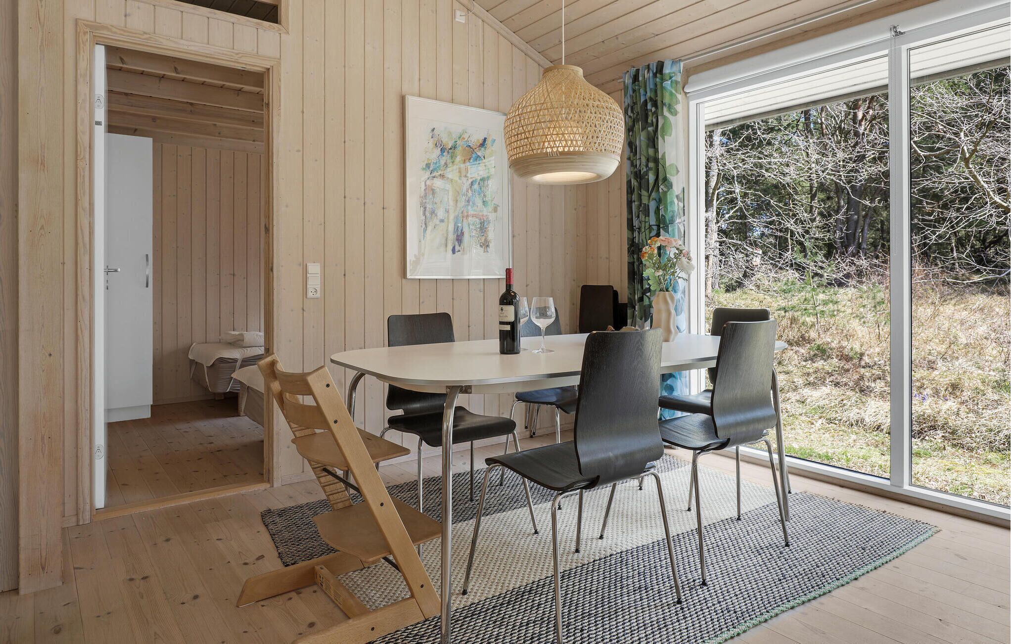Un coin repas avec une table ronde, des chaises noires, un luminaire suspendu et une grande fenêtre donnant sur la forêt.