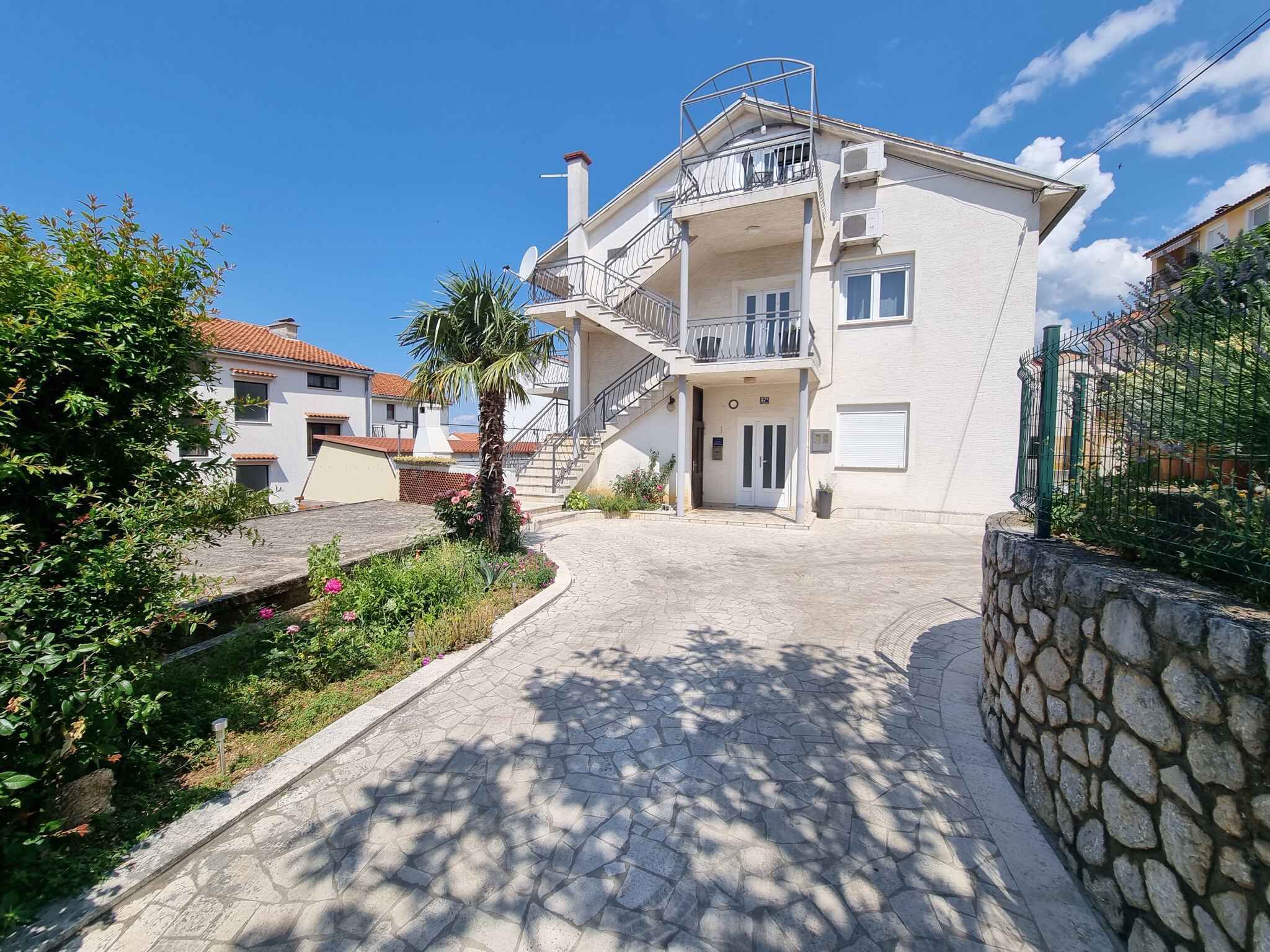 A two-story building with a balcony, a stone wall, and a paved pathway leading to the entrance.
