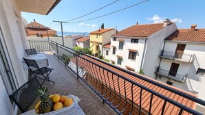 Un balcon avec une table dressée pour deux personnes, un panier de fruits contenant des ananas et des oranges, et une vue sur des bâtiments aux toits de tuiles.