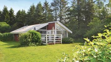 Une petite cabane à un seul étage, avec un toit rouge et des murs blancs, entourée d’une végétation luxuriante et de grands arbres.