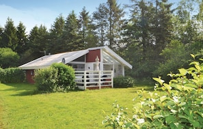 Une petite cabane à un seul étage, avec un toit rouge et des murs blancs, entourée d’une végétation luxuriante et de grands arbres.