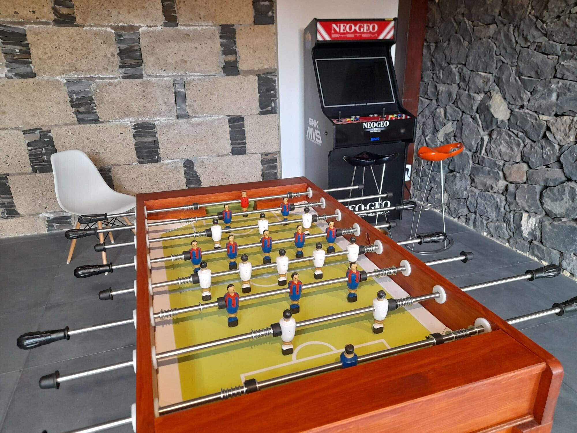 Ein Kicker-Tisch vor einer Steinwand, im Hintergrund steht ein Neo-GEO Arcade-Automaten.