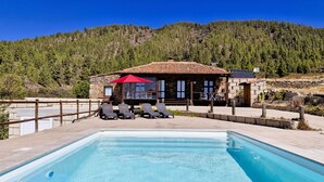 A pool area with lounge chairs and a red umbrella, a stone building with a tiled roof, and a forested hillside in the background. - Casa Rural Altos Cruz de Tea (Granadilla de Abona)